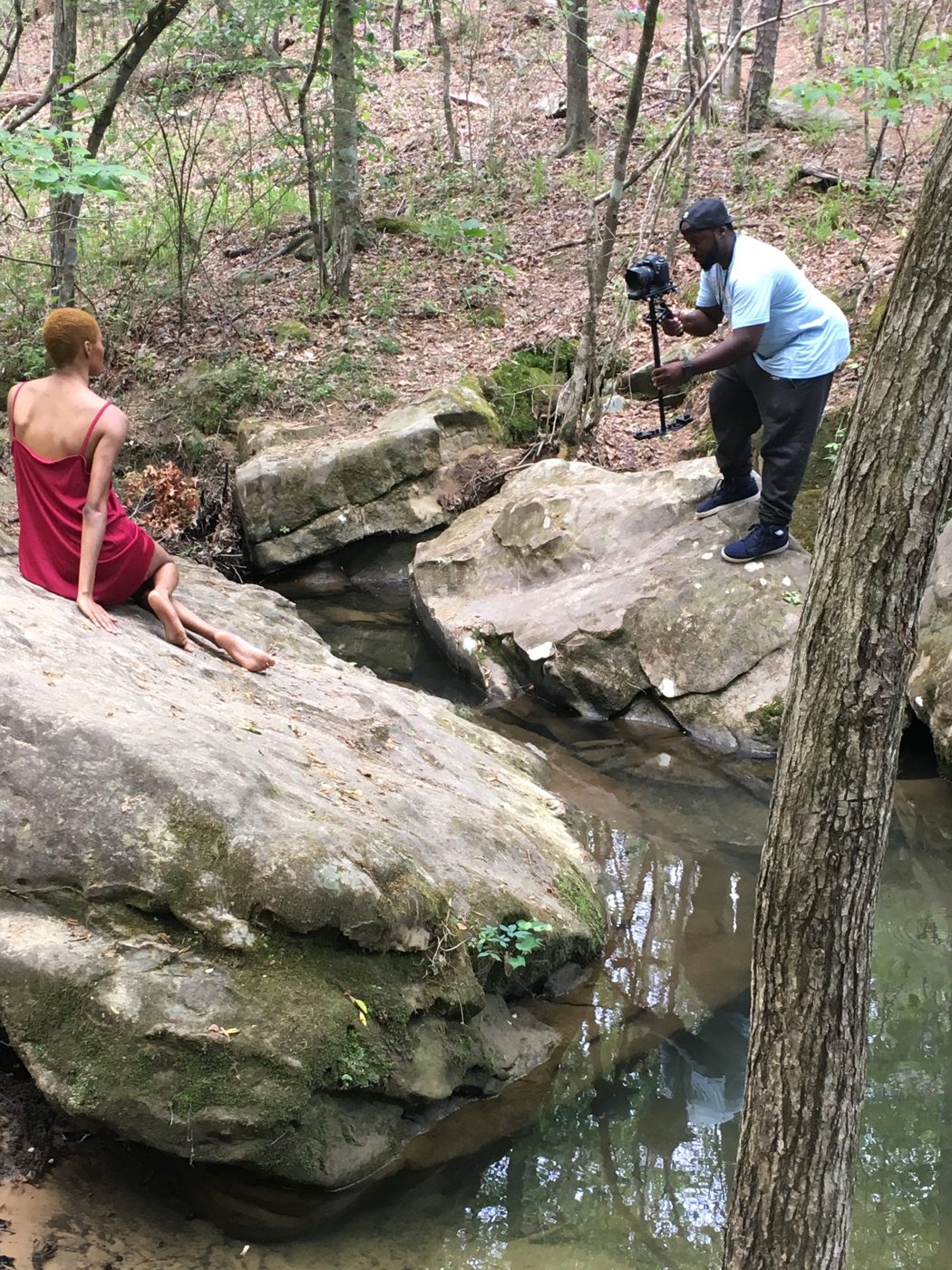 A videographer with a stabilizer films the model seated on a rock beside a creek