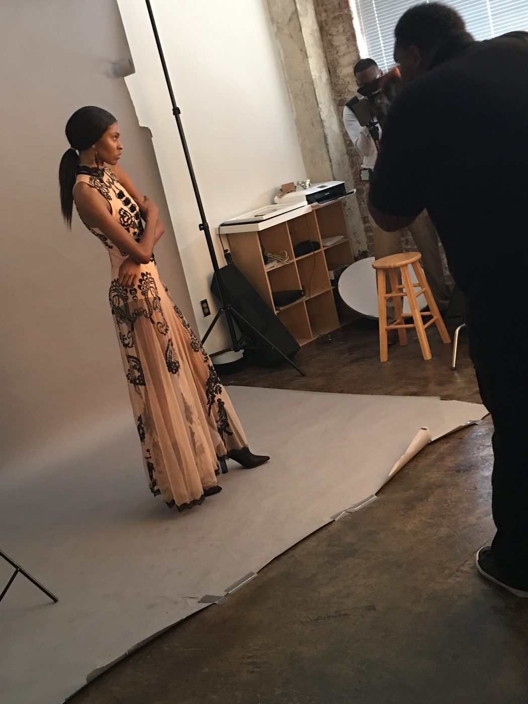 The model holds a pose in a lace gown on a paper backdrop while photographers work in a loft studio
