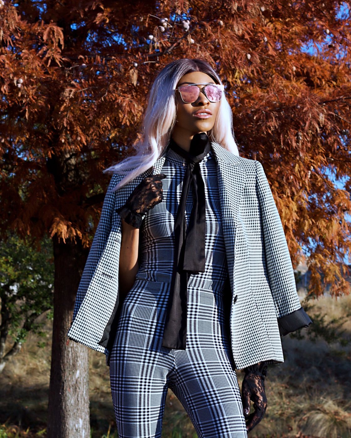 Infinity Jasele in a houndstooth blazer, plaid jumpsuit and pink mirrored sunglasses beneath autumn trees